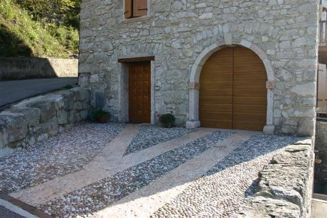 Facciata di casa in pietra con portoni in legno e vialetto in ciottoli.