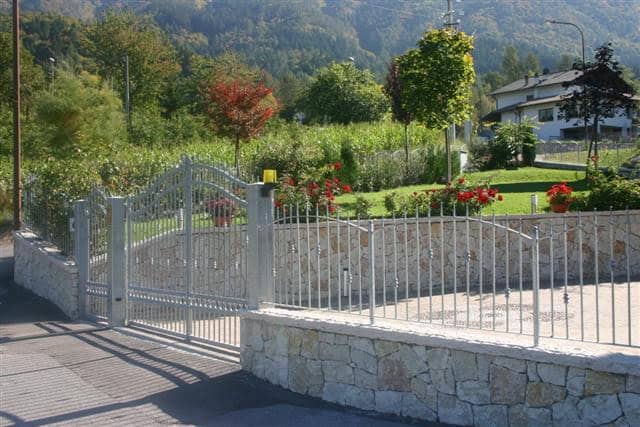 Cancello in ferro battuto con recinzione in pietra, giardino curato con alberi e fiori.