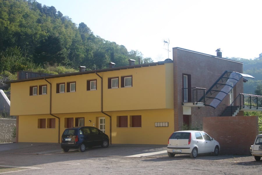 Edificio residenziale giallo con parcheggi e vista su colline verdi.