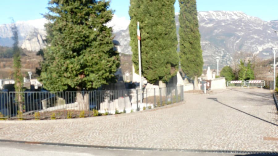 Cimitero di Lasino con monumenti e alberi, paesaggio montano sullo sfondo.