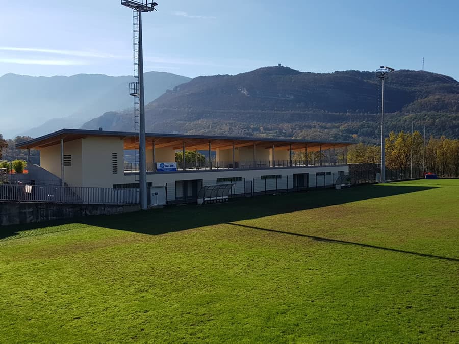 Stadio di calcio con tribune e luci, montagna sullo sfondo, in Italia.