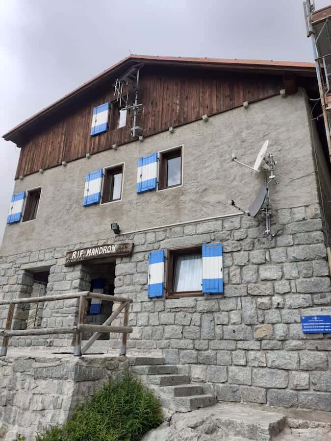 Costruzioni Dallapè Srl, Rifugio Mandron, edificio in pietra e legno con finestre con persiane blu.