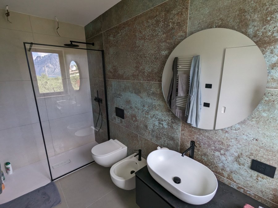 Bagno moderno con lavabo, bidet e doccia a vetro, vista su parete decorativa rustica.